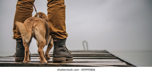 Man In Boots Protecting His Dog From Rain And Standing On Old Wooden Deck