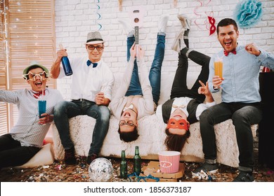 A Man With A Bottle Of Alcohol Sits On The Couch And Smiles. Two Men Turn Upside Down And Have Fun At A Gay Party. 