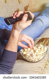 A Man With A Bowl Of Popcorn And A Remote Control In His Hand Looks At The TV On The Sofa