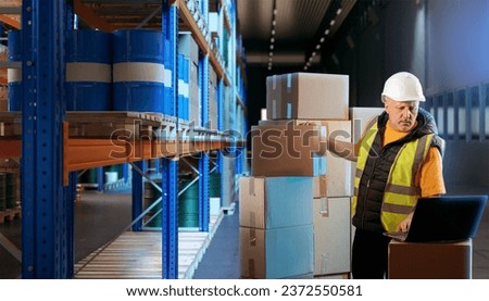 Man with boxes in warehouse. Loader keeps records of goods in storage. Warehouse manager with laptop. Guy works in fulfillment center. Racks with barrels near loader. Worker in warehouse hangar