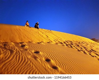 Man and boy, with not recognizable faces, seat at the top of orange sand dune with deep blue sky behind