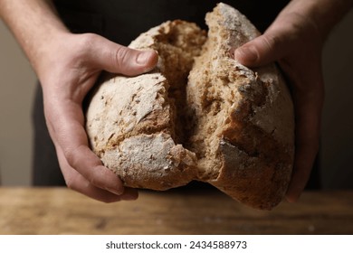 Man breaking loaf of fresh bread at wooden table, closeup