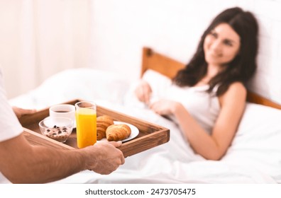 A man is bringing a breakfast tray to a woman in bed. The tray contains a cup of coffee, a glass of orange juice, a bowl of cereal, and two croissants. The woman is smiling and looking at the tray. - Powered by Shutterstock