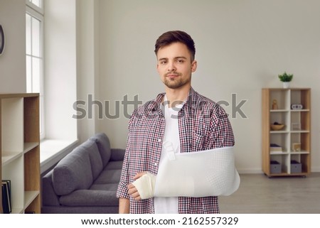 Man with broken arm is rehabilitated with help of medical devices to support his arm. Portrait of young man with white bandage on broken arm standing in living room at home and not smiling.