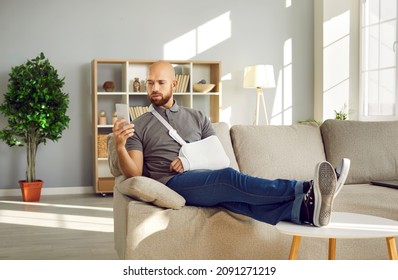 Man With Broken Arm Using Mobile Phone. Male Patient Wearing Medical Support Brace And Sling Bandage Immobilizer On His Elbow That Was Injured In Car Crash Relaxing On Sofa At Home And Calling Doctor