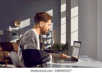 Man with broken arm working on laptop computer in office. Side view shot of businessman wearing arm splint sitting at desk in front of window working distantly at home - Shutterstock ID 2284463317