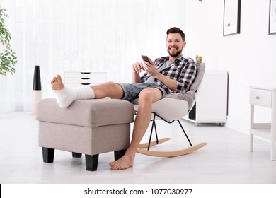 Man With Broken Leg In Cast Using Mobile Phone While Sitting In Armchair At Home