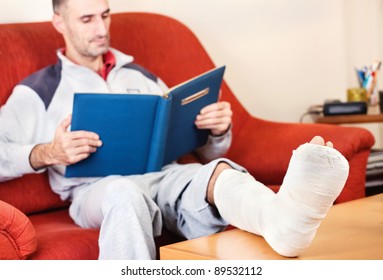 Man With A Broken Leg On A Sofa At Home  Reading Book; Focus On Cast