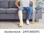 A man with a broken leg sits on the sofa in the living room, close-up. Concept injury, bone fracture, treatment and rehabilitation at home, accident. Use of crutches, medical care, traumatology