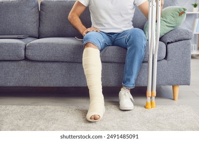 A man with a broken leg sits on the sofa in the living room, close-up. Concept injury, bone fracture, treatment and rehabilitation at home, accident. Use of crutches, medical care, traumatology