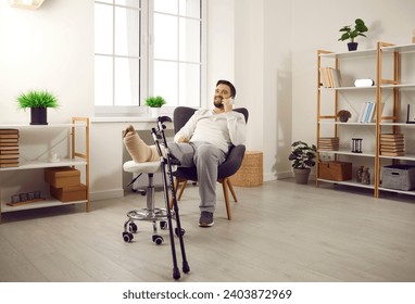 Hombre con la pierna rota sentado en un sillón hablando por teléfono. Un hombre guapo y alegre con la pierna enchapada y muletas recuperándose en casa. Fractura ósea, traumatología, lesión física