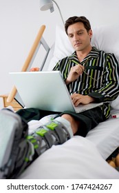 Man With Broken Leg Typing On Laptop In Hospital Bed