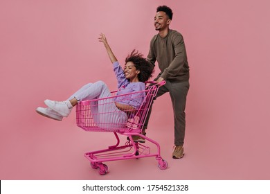 Man in brown outfit riding girlfriend in shopping cart on isolated. Portrait of guy and teen curly girl in purple outfit having fun on pink background
