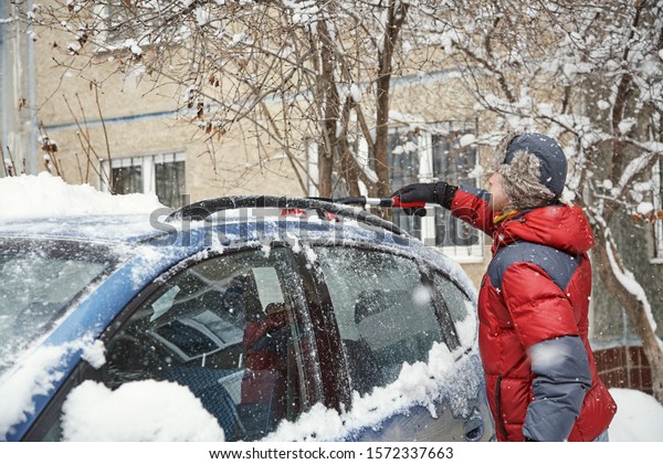 a man brushing the snow off the windshield of\
his car. use of an auto in\
winter