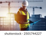 Man builder. Engineer with laptop. Builder near silhouettes unfinished buildings. Man architect stands with computer in hands. Builder in yellow vest and helmet. Architect against sunset background