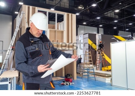 Man builder exhibition pavilions. Construction wooden structures inside hangar. Preparation pavilions at exhibition. Man with tablet and papers. Builder in helmet at production exhibition structures