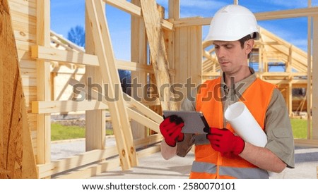 Man builder holding electronic tablet. Builder near frame of wooden house. Arhitect man. Erection of cottages made of timber. Builder in orange vest. Electronic construction documentation concept