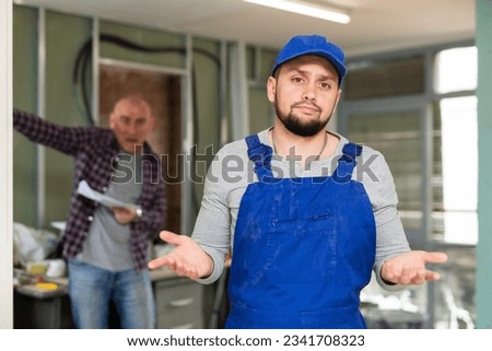 Man builder making helpless gesture while construction manager standing in background and shouting at him.