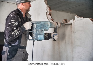 Man Builder In Workwear Drilling Wall With Electric Hammer Drill. Male Worker Using Electric Drill While Breaking Wall In Apartment Under Renovation. Demolishing And Construction Destroying Concept.