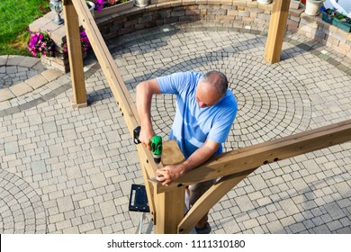 Man Building A Wooden Gazebo On A Brick Patio With Ornamental Paving Viewed From Above As He Stands On A Ladder Screwing Together Beams