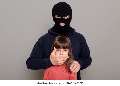 Man burglar in balaclava kidnapped little preschooler girl, holds her hostage, covers her mouth with his hands, asks for ransom for kidnapped child, posing isolated over gray background.