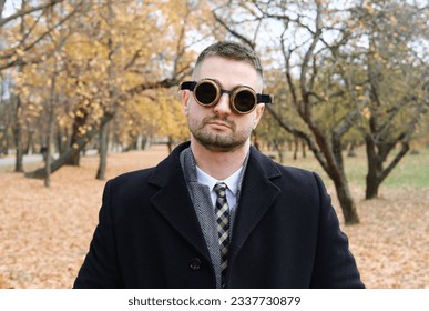 A man in a business suit, coat and steampunk goggles poses in an autumn park. Horizontal photo
