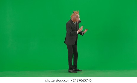 A man in a business suit with a horse head mask on a green studio background. A businessman is talking on a smartphone, gets angry and shouts into the phone. Hard office work concept. - Powered by Shutterstock