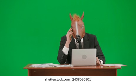 Man in business suit with horse head mask on studio green background. Businessman sitting at desk and talking on smartphone. Heavy office work concept. - Powered by Shutterstock