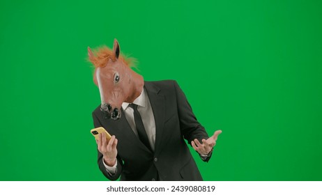 A man in a business suit with a horse head mask on a green studio background. A businessman is looking at something on his smartphone. Hard office work concept. - Powered by Shutterstock