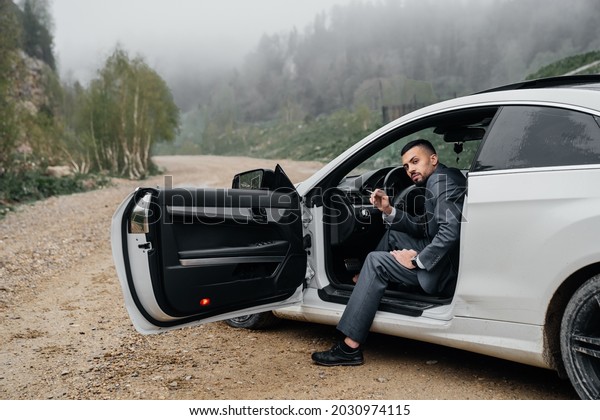 a man in a business suit sits behind the\
wheel of a car with the door open and smokes a cigarette. bad habit\
and dependence on nicotine. auto\
travel.