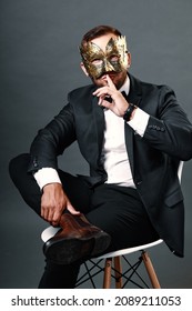 Man in business suit sits on chair wearing carnival mask on his face. Studio portrait on gray background. Young guy of 25-30 years hides his face under mask. Cunning and deceitful businessman