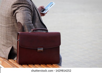 Man In A Business Suit Sitting With A Leather Briefcase On A Bench And Using Smartphone. Concept For Businessman, Official, Government Employee