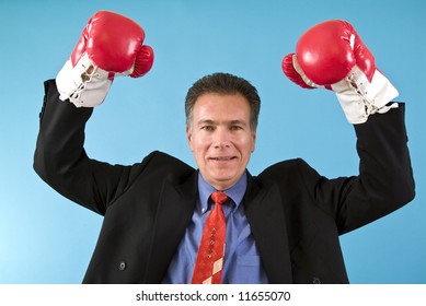 A man in a business suit wearing boxing gloves in a posture of triumph or winner.