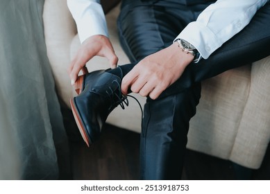 A man, a businessman on the wooden floor, ties his shoelaces with his hands on shiny black leather shoes. Wedding portrait of the groom in the morning. Preparation for work. - Powered by Shutterstock