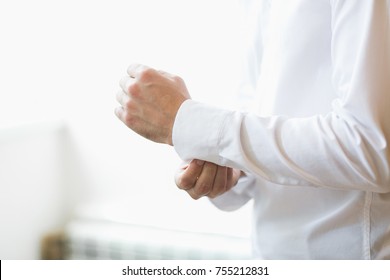 Man Buttons Cuff Link On Cuffs Sleeves Luxury White Shirt. Close Up Of Man Hand Wears White Shirt And Cufflinks. Groom Fixes Cufflinks Standing On White Background