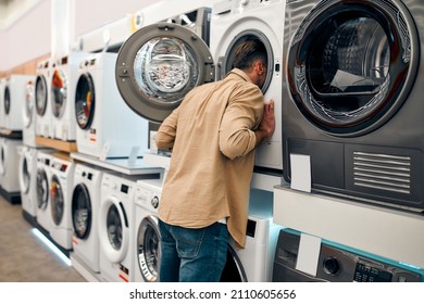 A Man Buying A Washing Machine In A Home Appliances And Electronics Store, Funny Sticking His Head Inside The Washing Machine, Looking Inside.