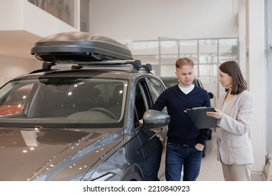 Man Buys A Car At A Car Dealership. A Female Salesperson And Car Rental Helps With The Purchase. Signing A Trade-in Contract And Handing Over Keys, Shaking Hands. A Successful Man Chooses A New Car