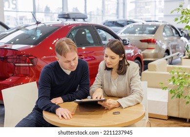 Man Buys A Car At A Car Dealership. A Female Salesperson And Car Rental Helps With The Purchase. Signing A Trade-in Contract And Handing Over Keys, Shaking Hands. A Successful Man Chooses A New Car