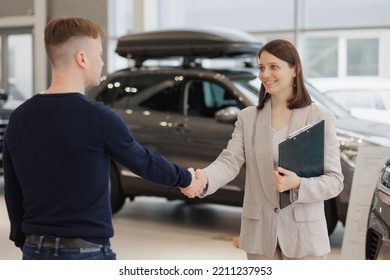 Man Buys A Car At A Car Dealership. A Female Salesperson And Car Rental Helps With The Purchase. Signing A Trade-in Contract And Handing Over Keys, Shaking Hands. A Successful Man Chooses A New Car