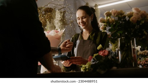 Man buys spring bouquet in flower shop and pays for purchase with contactless payment using smart watch. Female florist sells fresh flowers to male customer. Floristry and entrepreneurship concept. - Powered by Shutterstock