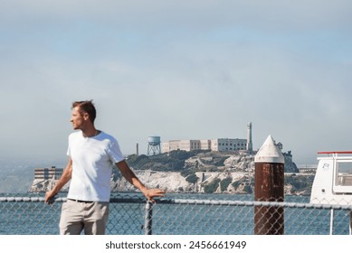 Man by fence looks towards water, Alcatraz Island in view. Famous structure and lighthouse seen. Clear weather with light fog. Tourist spot in San Francisco Bay, visited by many. - Powered by Shutterstock