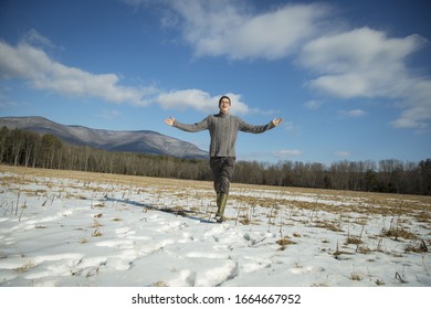 A Man In A Cable Knit Jumper And Muck Boots Standing With His Arms Stretched Out, In A Snowy Rural Landscape.