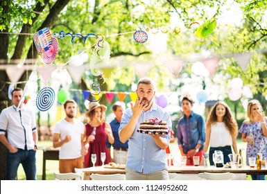 Man With A Cake On A Family Celebration Or A Garden Party Outside, Licking His Finger.