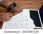 Man calculating taxes using smartphone and reviewing documents on wooden desk with laptop. Tax preparation and financial management concept 