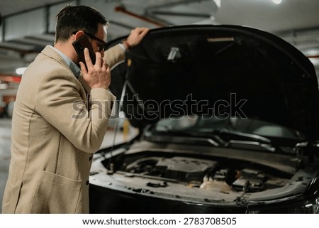Man calling for help with car breakdown, experiencing stress and unexpected problems in a garage setting with an open hood