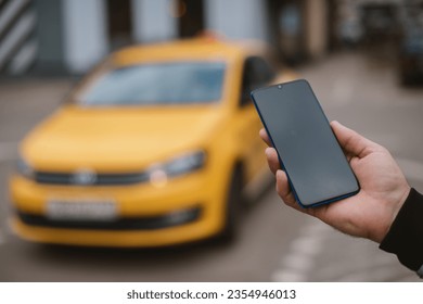 A man calls a taxi in the city through an app on his phone. Close-up. Yellow car on the background. The concept of urban transport. A guy with a phone in his hands is waiting for his taxi