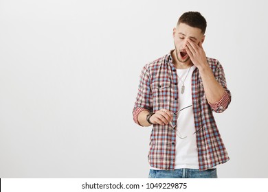 Man came from nightshift and needs some sleep. Portrait of cute unshaven male student yawning and rubbing eyes, being exhausted after work, holding glasses in hand. Guy is tired of doing homework