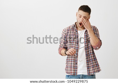 Man came from nightshift and needs some sleep. Portrait of cute unshaven male student yawning and rubbing eyes, being exhausted after work, holding glasses in hand. Guy is tired of doing homework