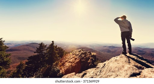 Man with a camera standing on at the edge of a cliff overlooking the mountains 
