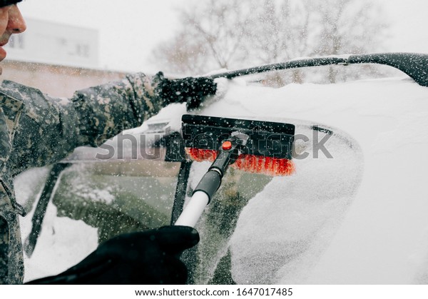 man in camouflage\
clothing without a face cleans the car from heavy snow with a black\
brush with a red pile. Cleaning the snow-covered glass of the car\
in bad weather.